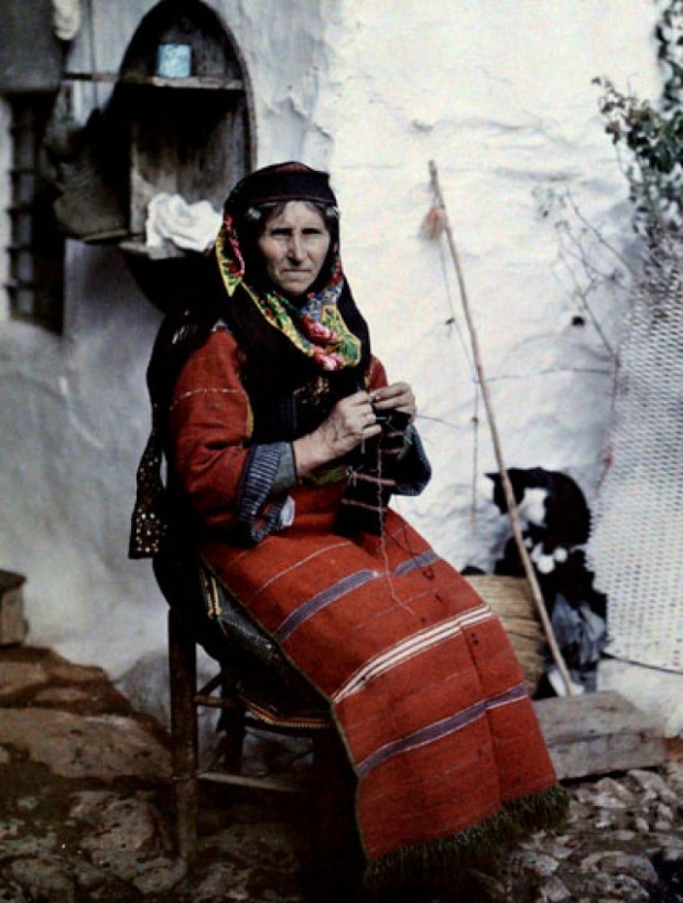 Μία γυναίκα με παραδοσιακή ελληνική ενδυμασία πλέκει έξω από το σπίτι της στο Αϊβαλί.